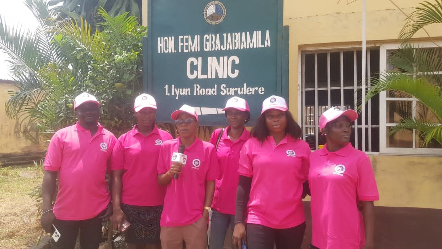 Seven WEWAD team members in branded pink polo shirts and caps stand outside the Hon. Femi Gbajabiamila Clinic on Iyun Road, Surulere, Lagos.